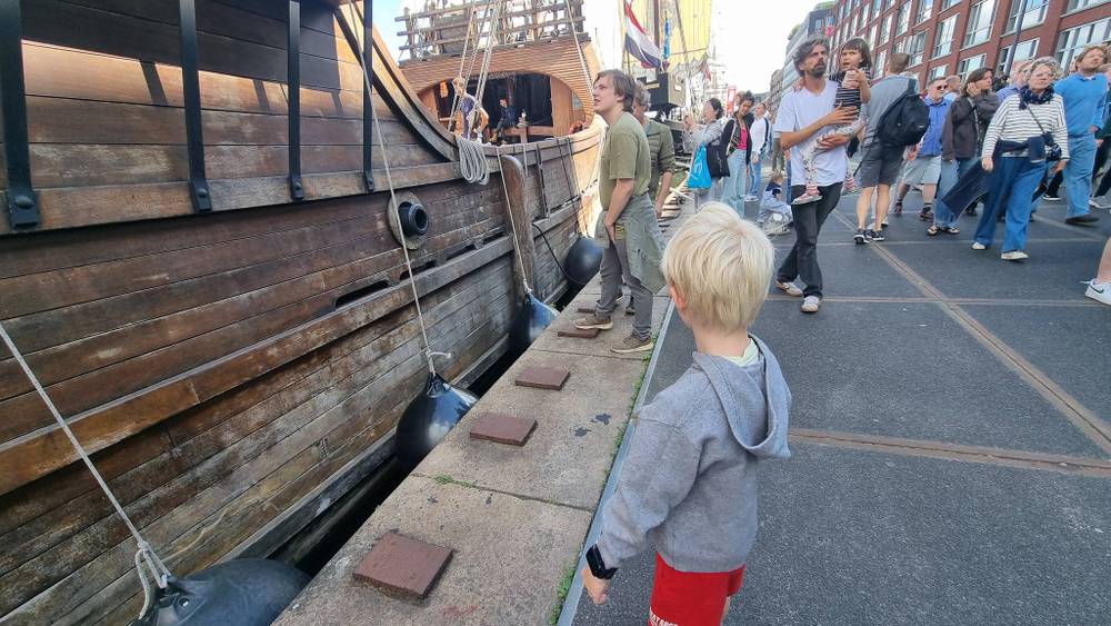 A child stands beside the wooden hull of a tall ship as people walk past.