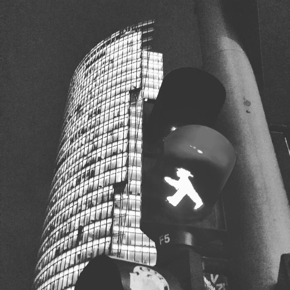 A black and white photo of a pedestrian traffic light showing a walking figure, with a building facade behind it.