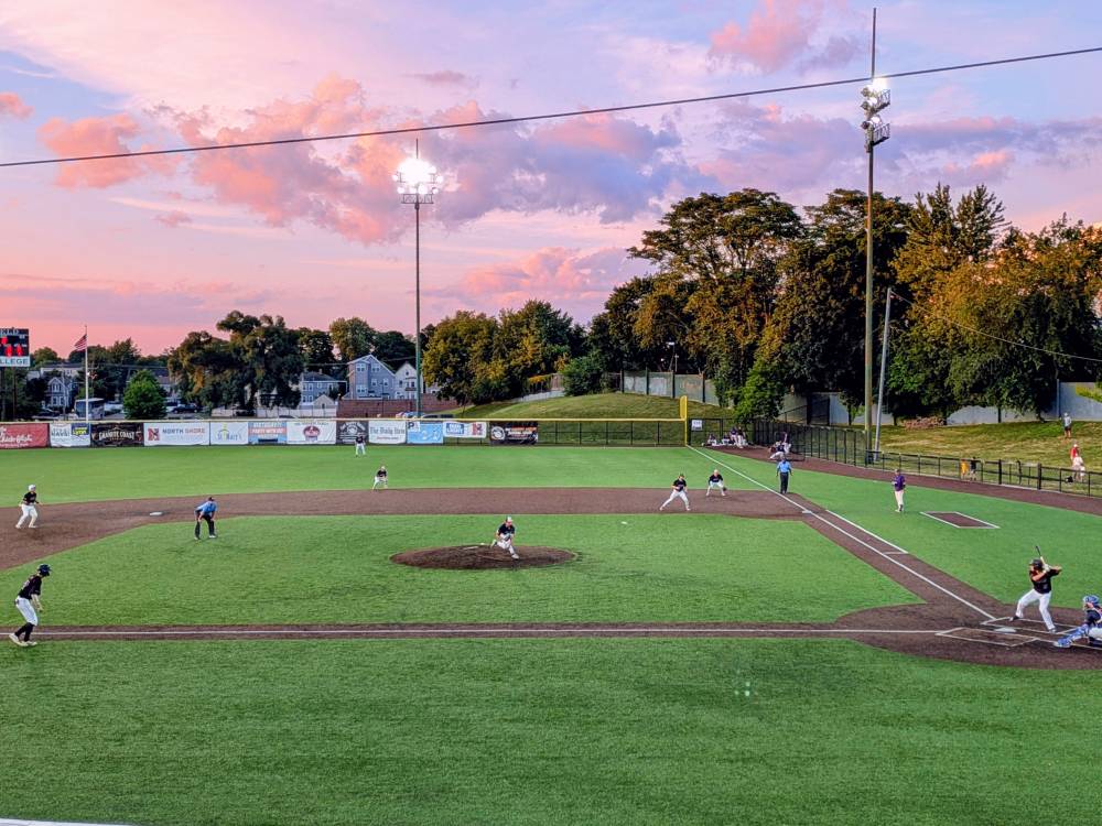 A pitcher for the North Shore Navigators throws a pitch, clouds are pink and blue in the sky and the lights glow above the field.