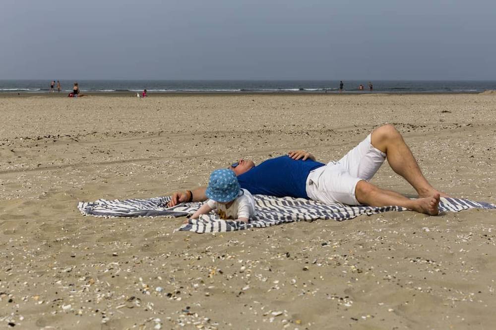 A man lies on a striped beach blanket beside a baby by the sea.