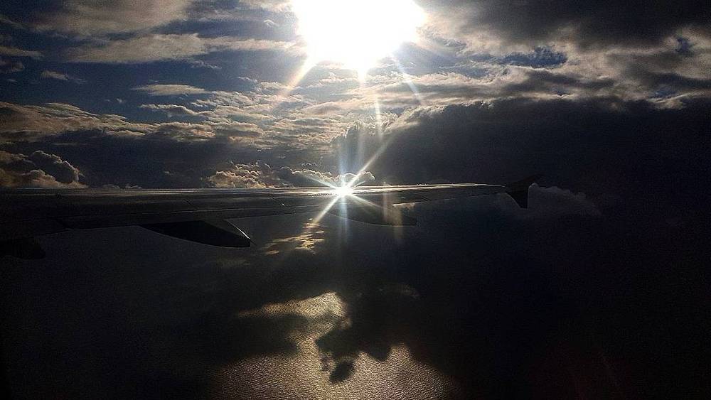 Dramatic sunburst through clouds seen from an airplane window, with a dark landscape below.