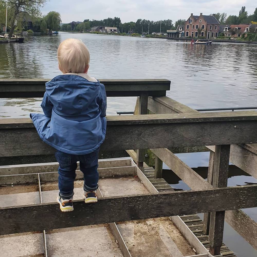A child in a blue jacket standing at a wooden railing looking out over a river with boats and houses.