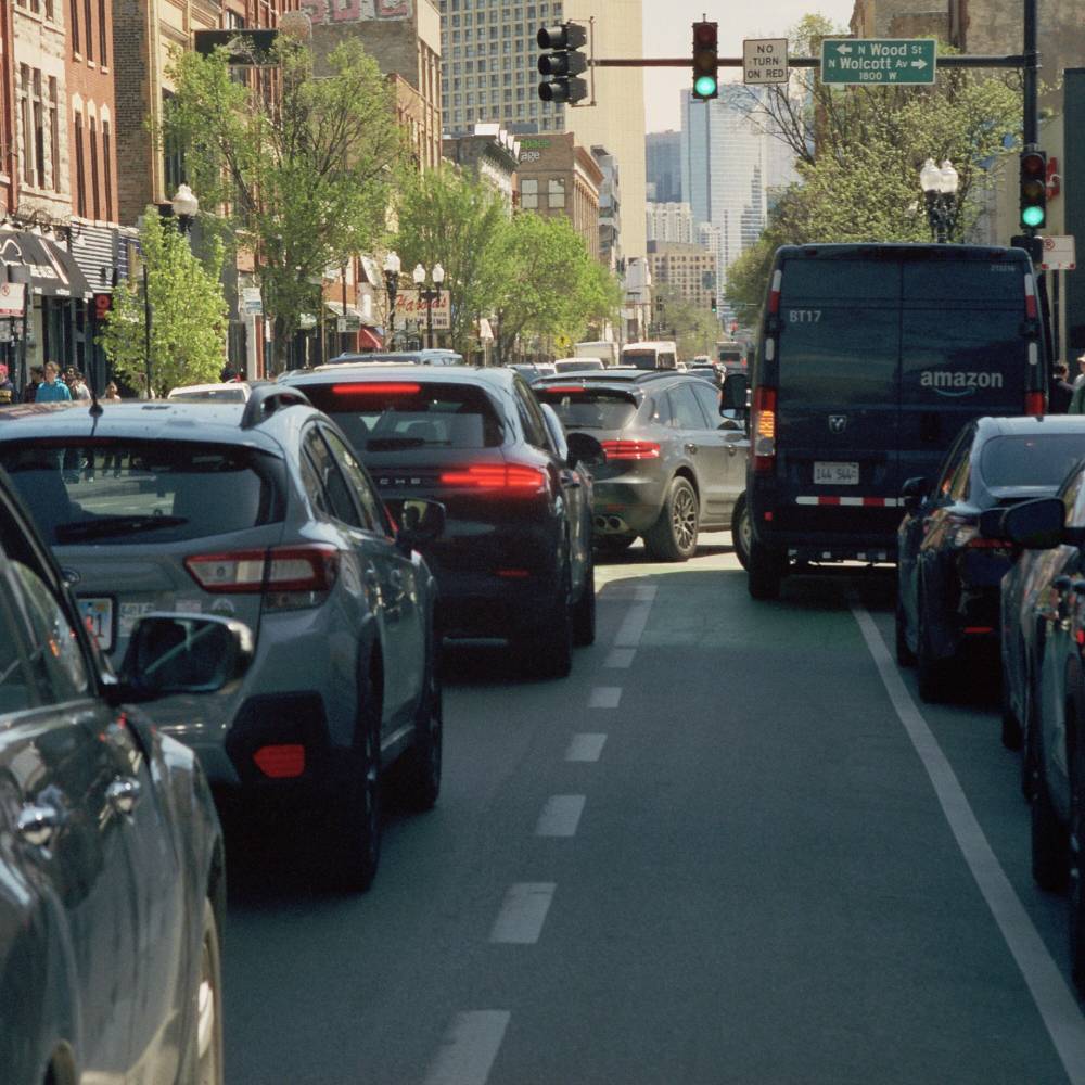 Heavy traffic on a Chicago street with a green-painted bike lane down the center, an Amazon delivery van on the right, and the downtown skyline visible ahead near Wood St and Wolcott Ave.