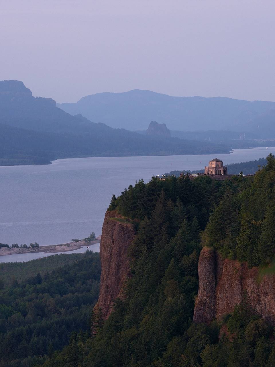 Vista House at Crown Point sits atop a basalt cliff overlooking the Columbia River Gorge at dusk, with Beacon Rock visible in the hazy blue distance and forested ridgelines receding into atmospheric haze.