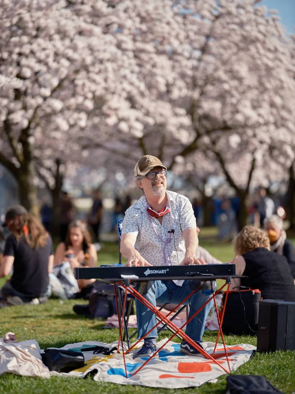 A bearded musician in glasses and tan cap sits cross-legged on a colorful blanket, fingers dancing across the keys of his portable keyboard as crowds gather beneath the ethereal pink bloom of cherry blossoms at Tom McCall Waterfront Park. The scene captures the quintessential Portland spring experience, where music and nature converge in perfect harmony. Soft afternoon light filters through the delicate sakura petals, creating a dreamlike backdrop for this impromptu street performance. Families and visitors scattered across the grass complete this snapshot of urban tranquility in Old Town Portland.