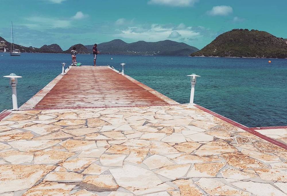A stone pier extending into the sea with hills in the background and two people at the end.