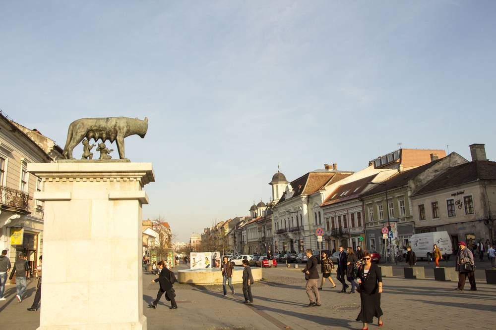 The wolf statue viewed from the square with pedestrians walking in the distance.