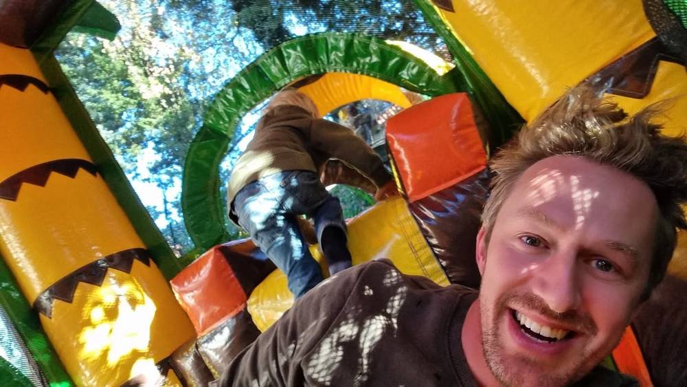 A man taking a selfie on a colorful bouncy castle while a child jumps in the background.