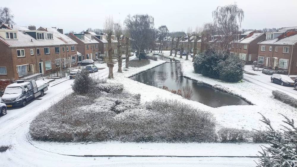 A snow-covered Dutch neighborhood with a canal, houses and parked cars.