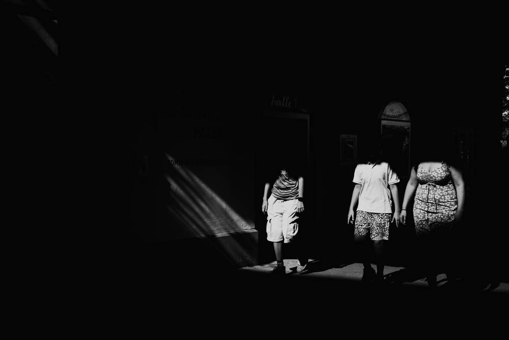 High-contrast black and white street photograph showing three headless silhouettes walking side by side through a beam of bright sunlight surrounded by deep shadows.