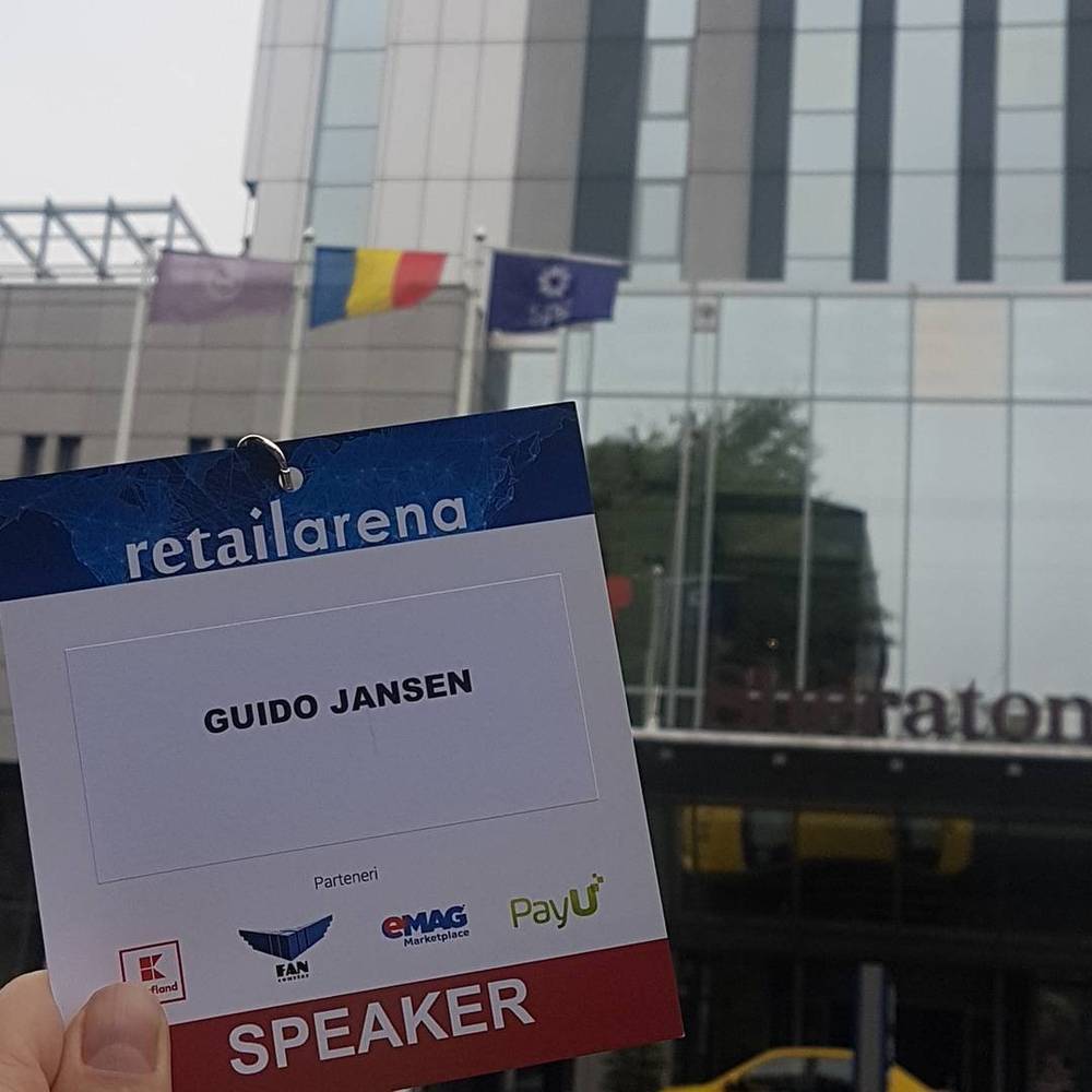 Guido Jansen's RetailArena speaker badge held up in front of a hotel building with flags, Bucharest.