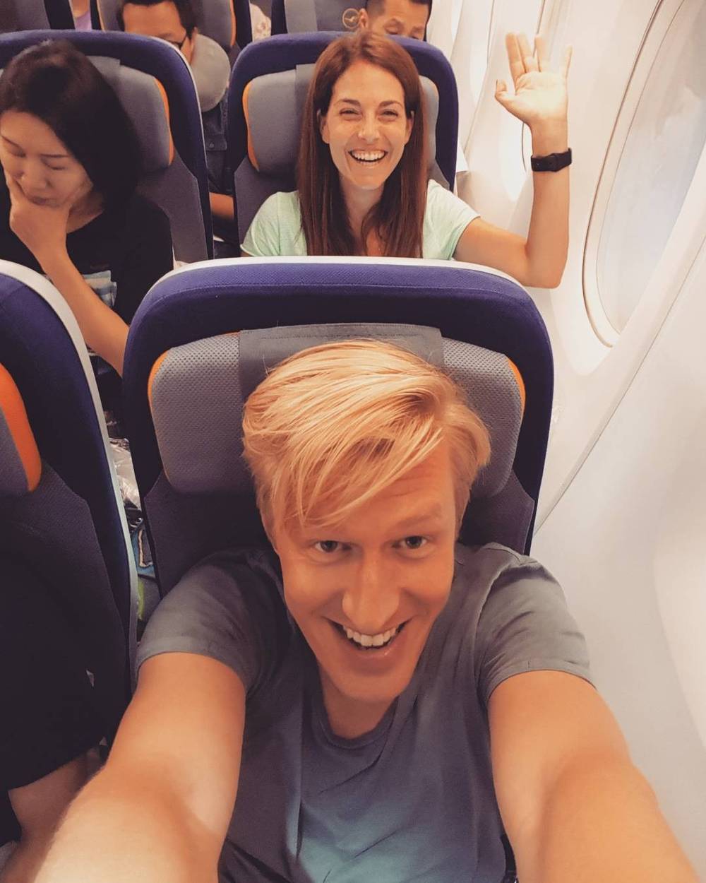 A smiling woman waving from her airplane seat, in a selfie with another passenger visible.