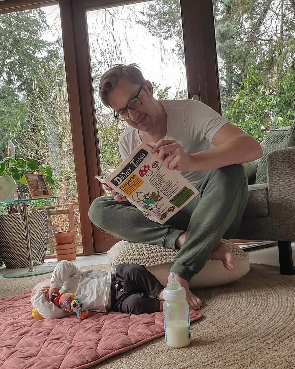 A man reads a children's book on the floor beside a baby lying nearby with a bottle.