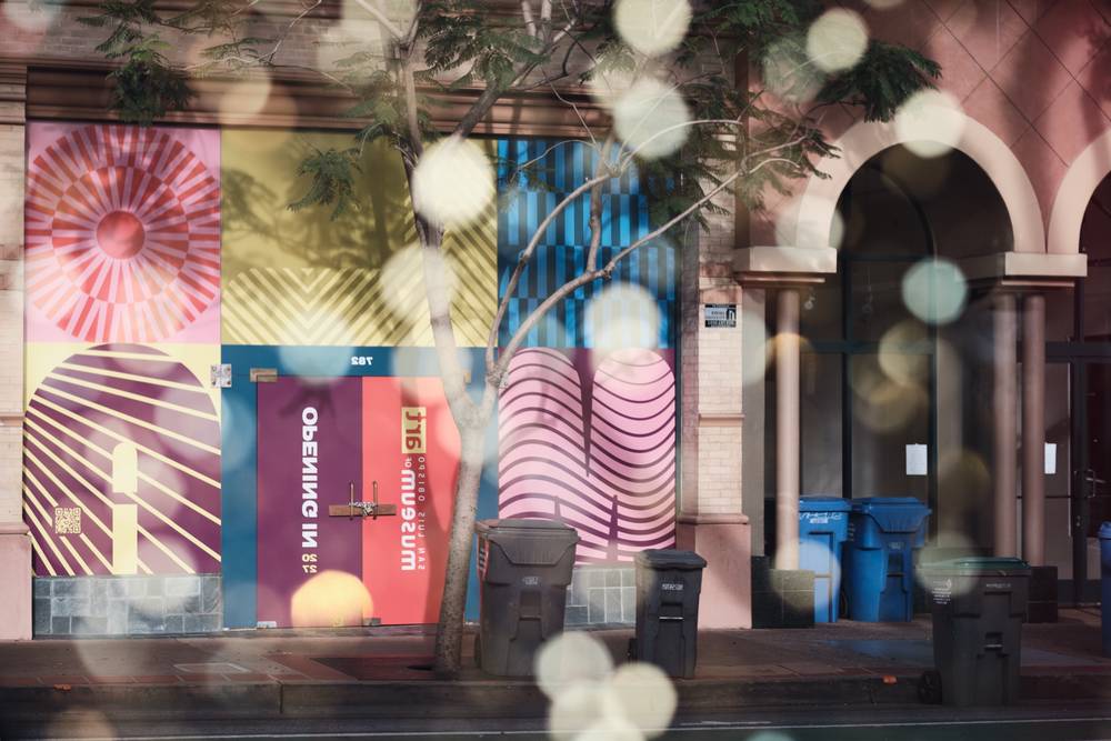 Photo of reflection of windows with tree shadows and bokeh balls covered in a wrap announcing art museum is moving in soon.