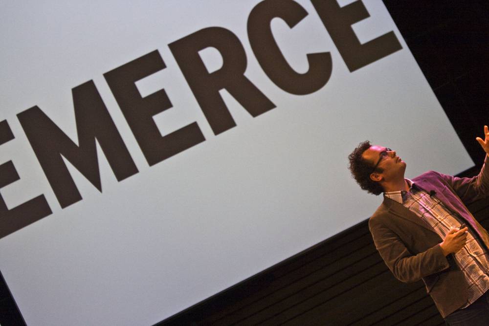 The speaker gestures energetically beside the large 'EMERCE' screen.