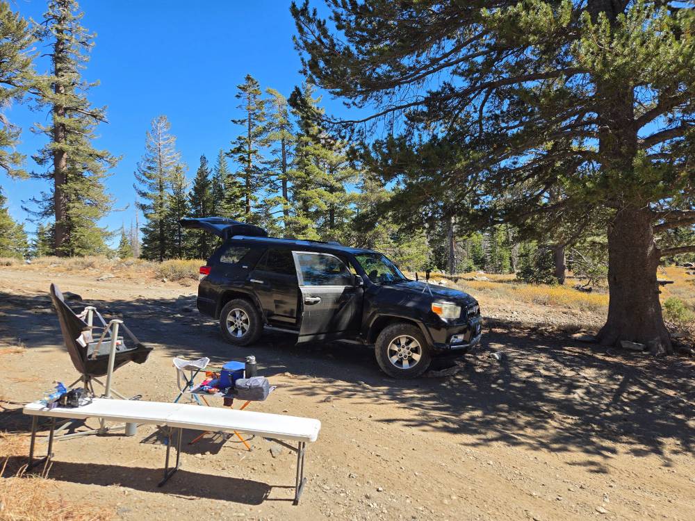 A black 4Runner with open doors is parked in the shade of a pine tree to the right. The open dirt clearing around the truck has a large swinging camp chair, a small folding chair serving as an ottoman, a narrow white folding bench, and a roll up camp table holding various lunch items. Tall green pine trees surround the clearing, with brilliant blue sky above.