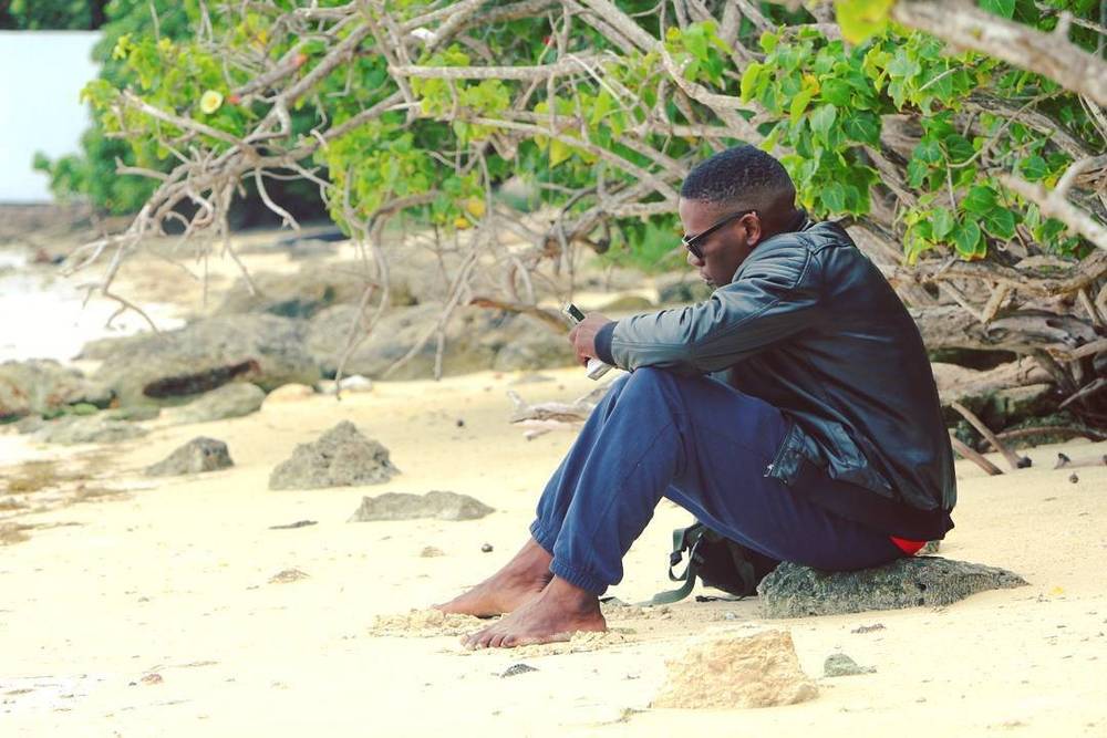 A man sitting barefoot on a beach under overhanging branches.