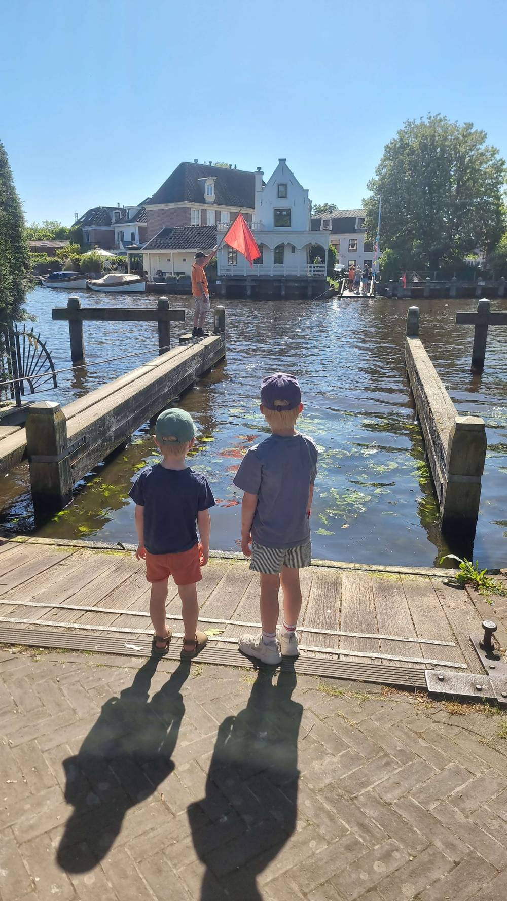 Two children stand on a wooden dock looking at the water, houses in the background.