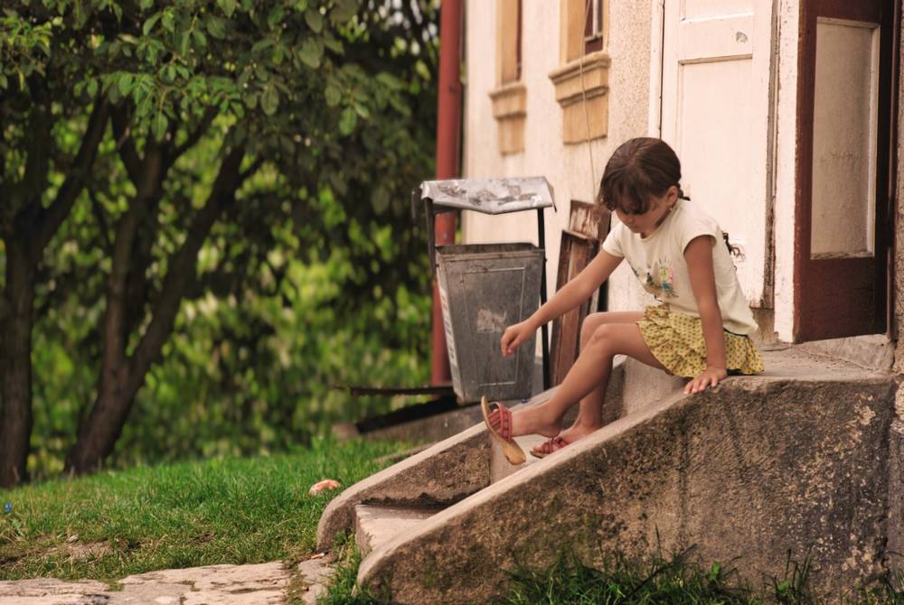 Una niña pequeña está sentada en los escalones de hormigón de la entrada de una casa, mirando hacia abajo. Viste una camiseta color crema, una falda amarilla con estampado floral y sandalias rojas, con un pie colgando. A su izquierda hay una papelera metálica con tapa y, más allá, un árbol de hojas verdes y hierba. La fachada de la casa es de color claro, con una puerta de marco marrón y ventanas.