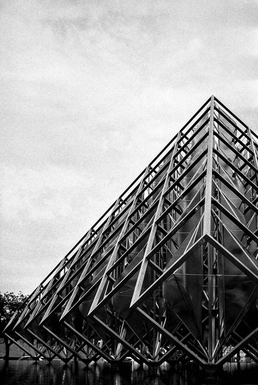 Black-and-white film photograph of Alejandro Otero’s Delta Solar in Washington, D.C. The geometric steel sculpture rises diagonally from a reflecting pool, its dense lattice of triangular beams and panels forming a sharp peak against a bright, cloudy sky. The low-angle composition leaves a broad expanse of textured sky above the dark, angular structure.