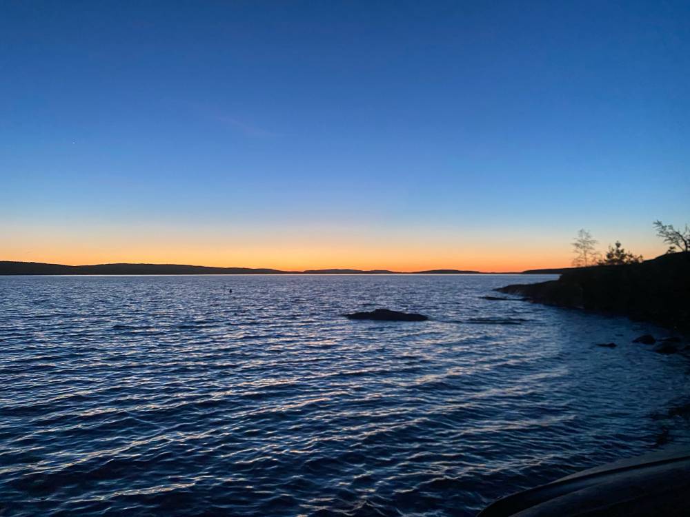 A wide lake at dusk, the horizon glowing orange and gold beneath a deep blue sky, silhouetted hills and pine trees framing the right edge.