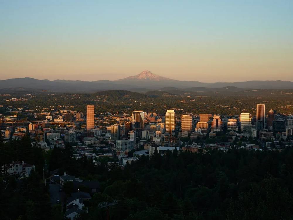 An elevated view of Portland, Oregon's downtown skyline at golden hour, with the snow-capped peak of Mount Hood visible in the distance beyond rolling hills. The city's high-rise buildings are illuminated by warm sunset light, while residential areas and dense forest cover the foreground and surrounding landscape.