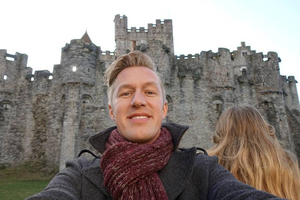 A man smiles for a selfie in front of a stone castle with a woman beside him.