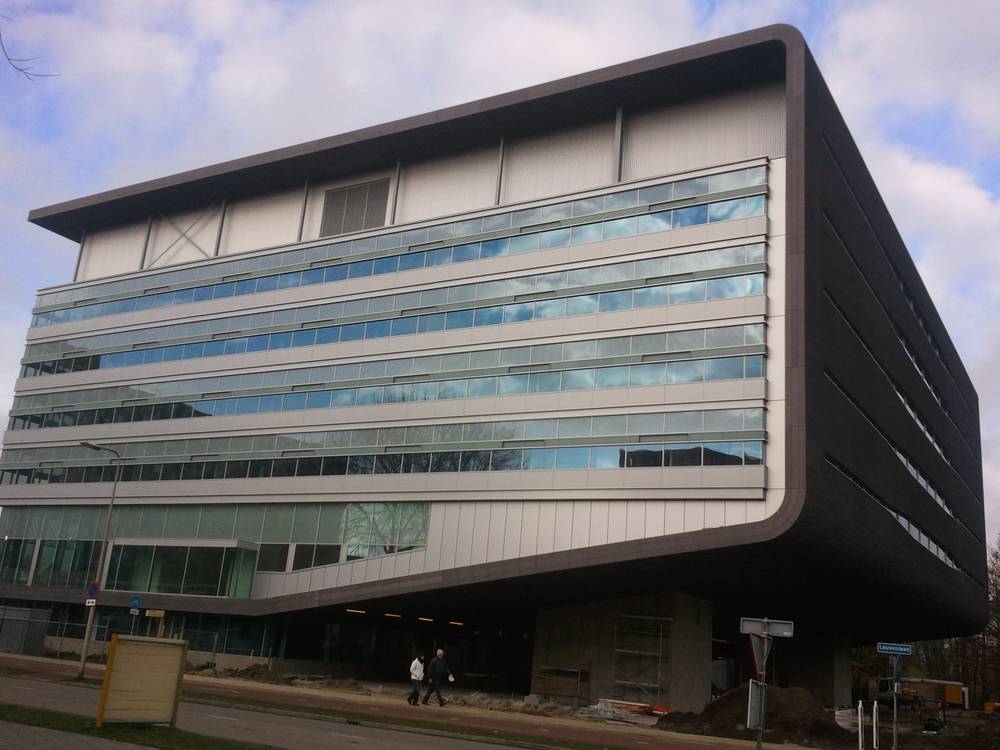 A modern dark office building with a cantilevered upper floor stands under a cloudy sky.