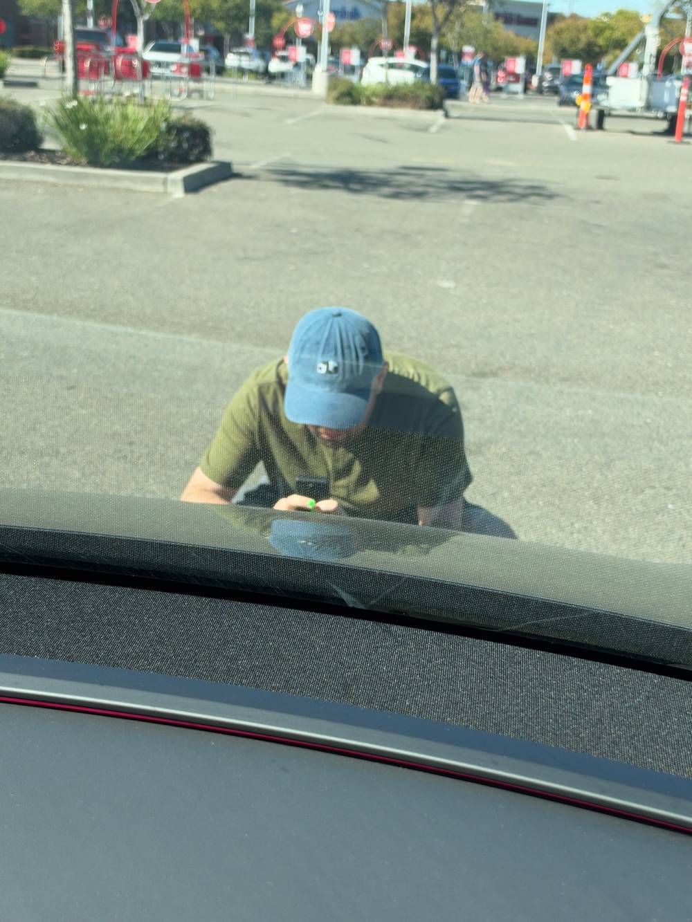 A person in a green shirt and blue cap leans toward a car windshield, holding a Game Boy Camera to photograph the license plate, seen through the glass with a parking lot and red signs in the background.