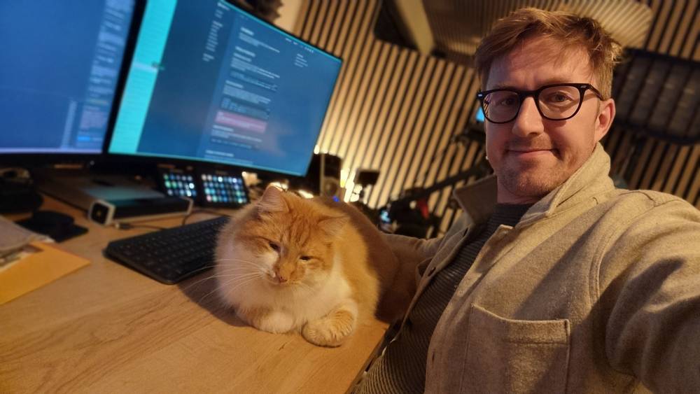 A man takes a selfie at his multi-monitor desk with a ginger cat sitting beside him.