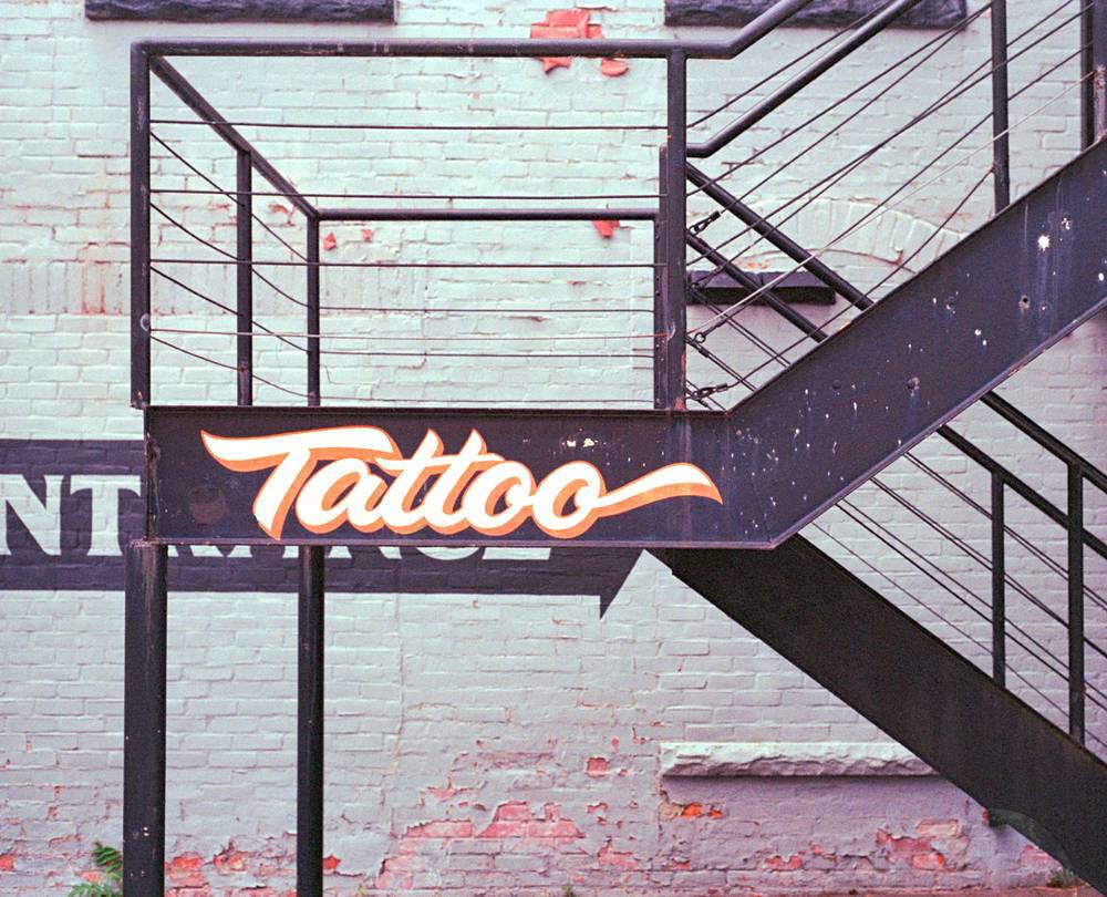 The word Tattoo is painted in orange script on the side of a black exterior metal staircase against a faded brick wall.
