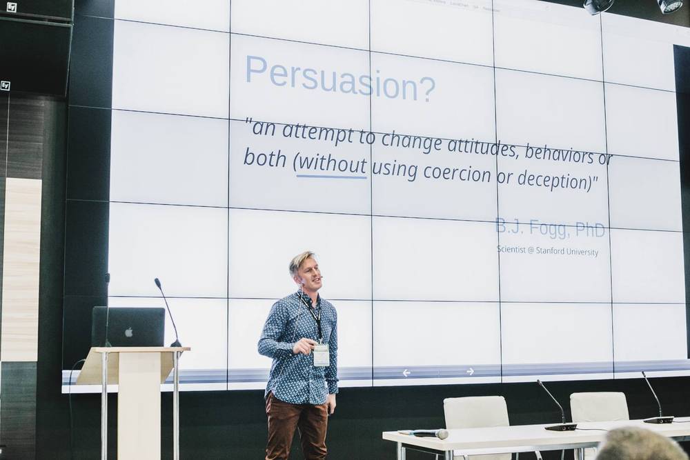A speaker stands beside a slide reading "Persuasion?" quoting B.J. Fogg during a conference talk.