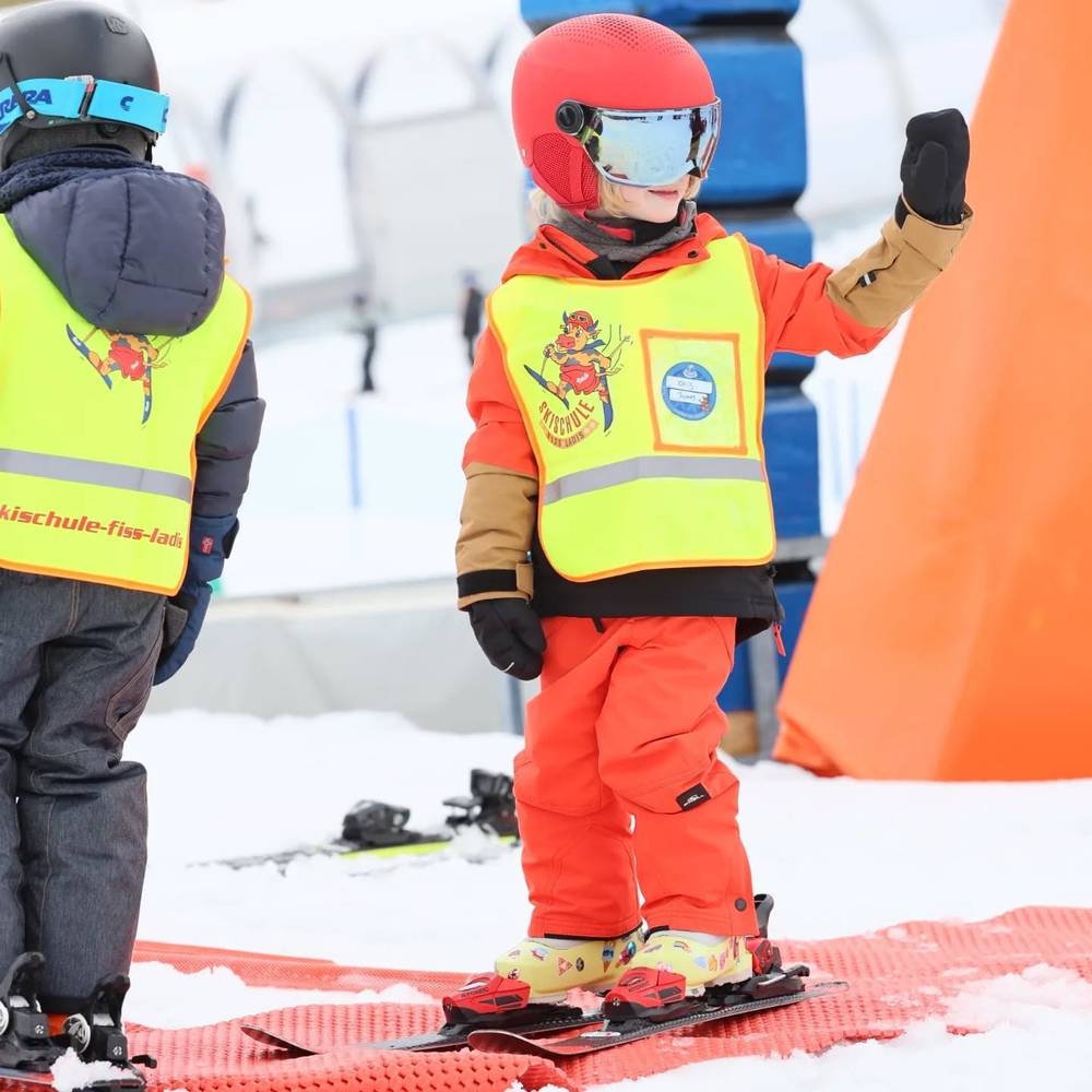 Two children in reflective ski-school vests standing together on a ski conveyor lift.