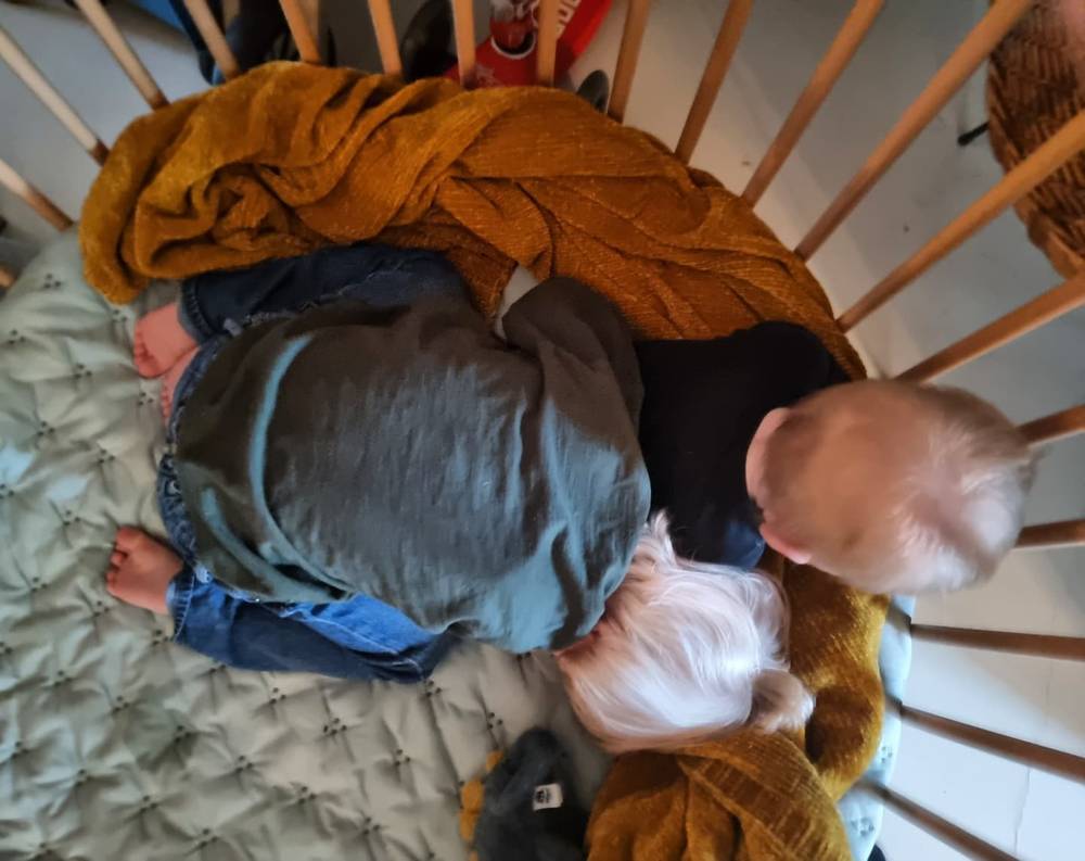 Two children lying together in a round crib, one wrapped in a mustard-colored blanket.