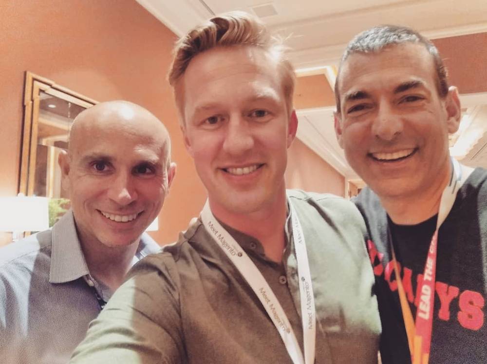 Three men smiling for a selfie indoors, wearing conference lanyards.