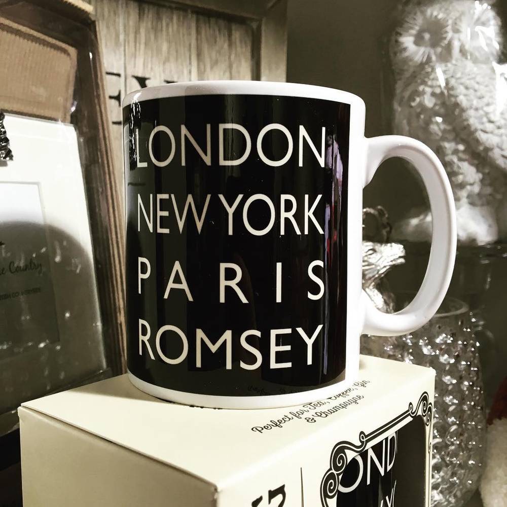 A mug printed with "London New York Paris Romsey" displayed among gift items and an owl figurine.