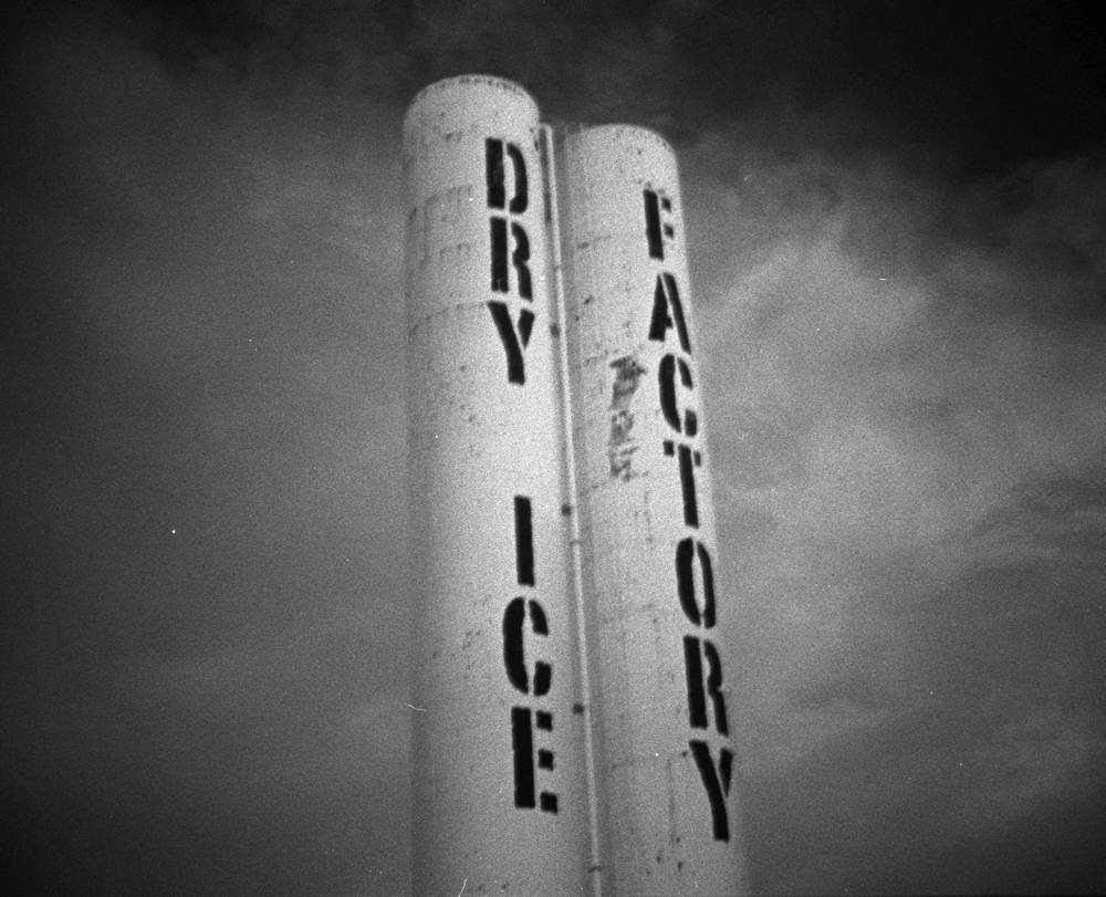 Two tall white industrial silos labeled DRY ICE FACTORY in stenciled black letters against a dark, cloudy sky in Denver's RiNo neighborhood.