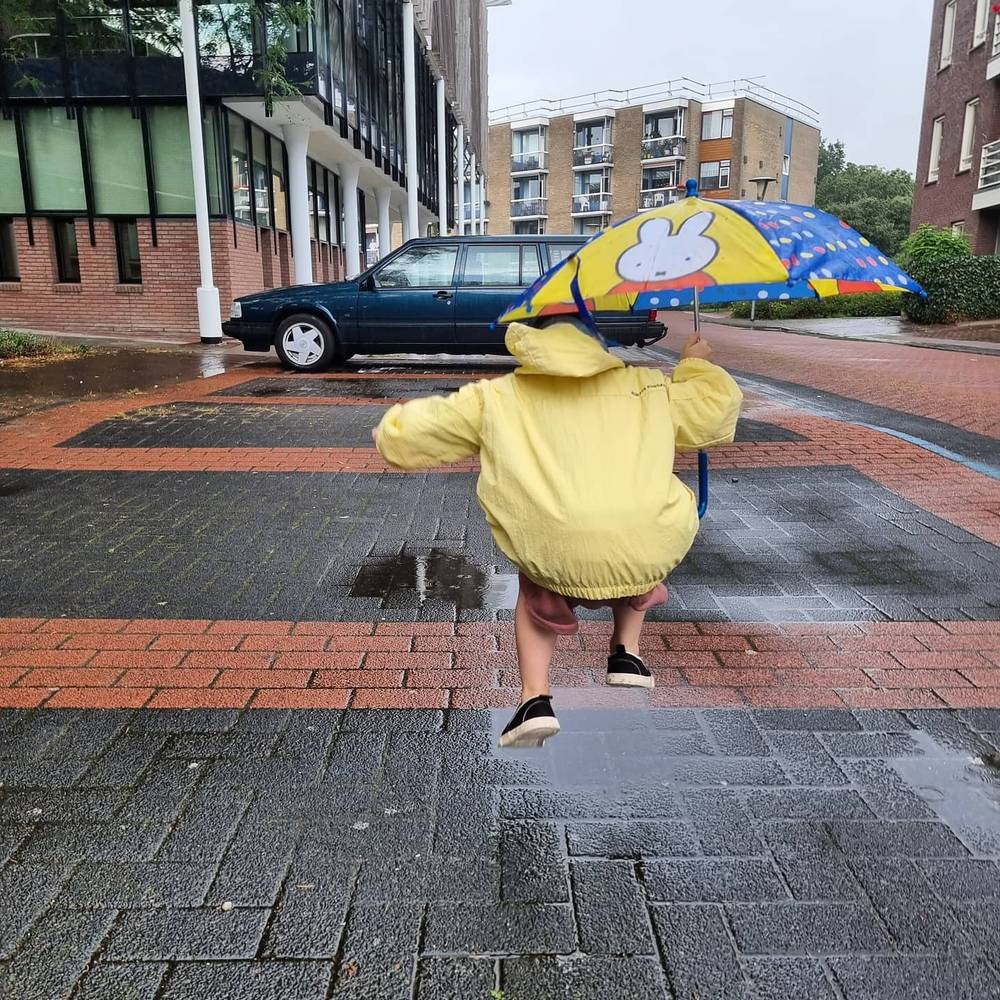 A child in a yellow raincoat running across a wet paved square, with a colorful Miffy-themed umbrella nearby.
