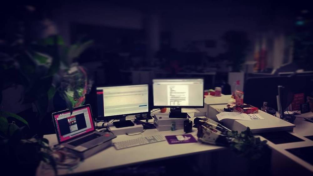 A dimly lit office desk with multiple computer monitors displaying work late at night.
