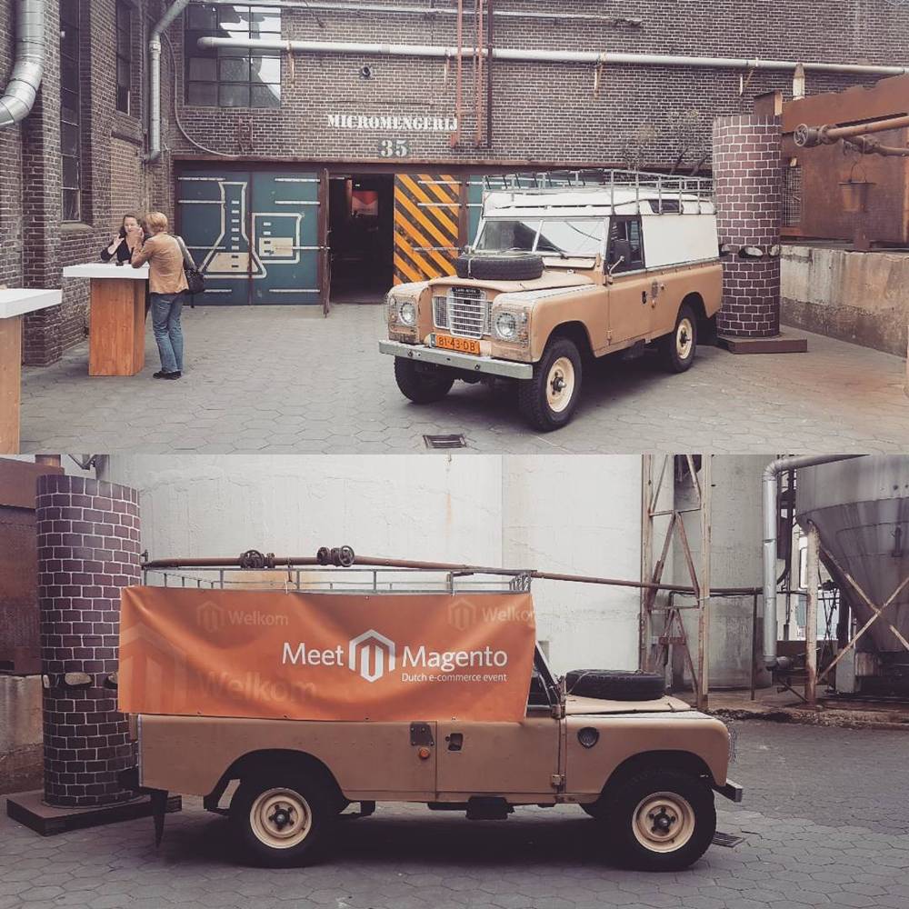 A vintage Land Rover displaying a Meet Magento welcome banner outside an event venue.