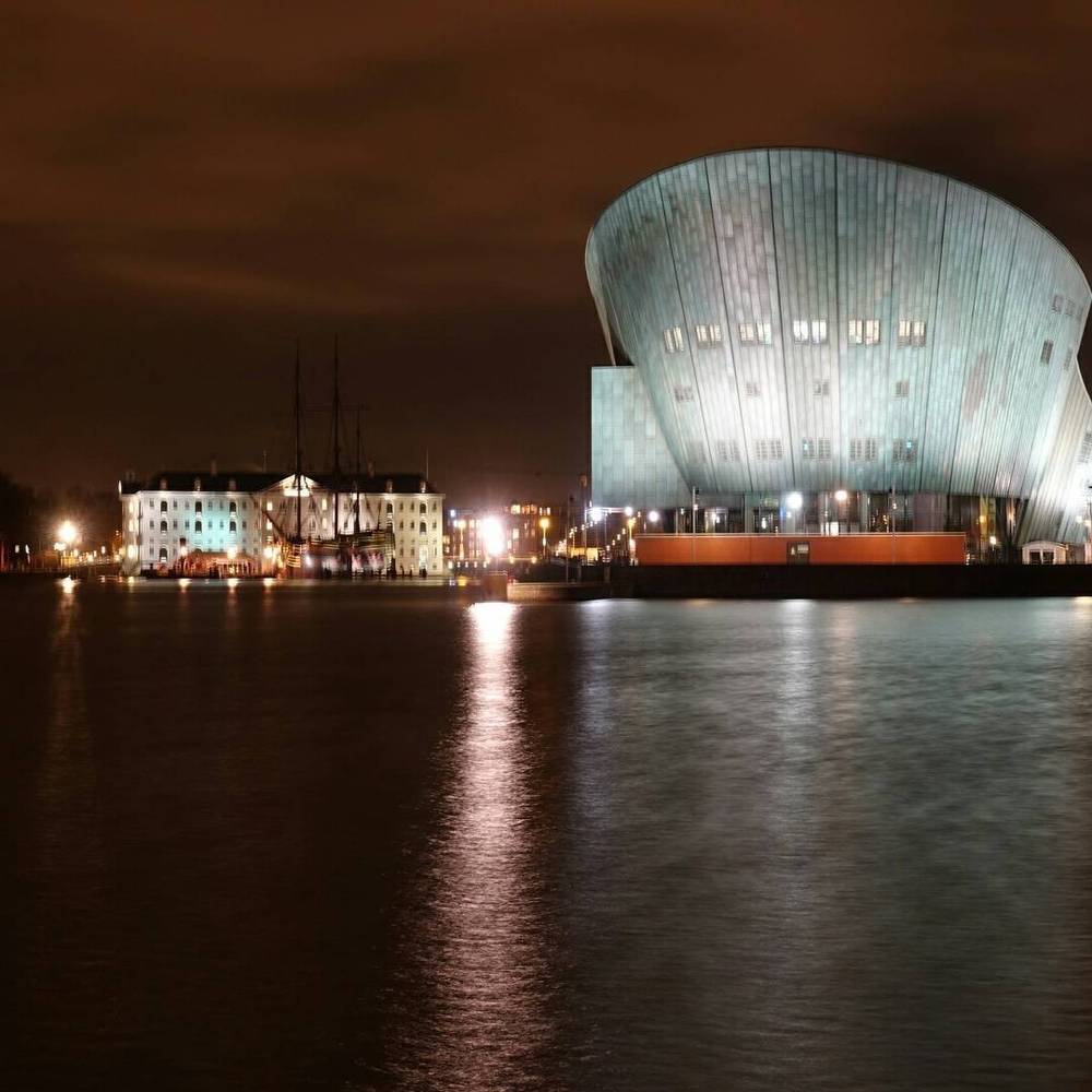The NEMO Science Museum building illuminated at night, reflected in the harbor water.