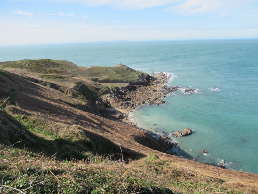 High-angle view of a rugged coastline featuring steep, grassy cliffs sloping down to a rocky shore and turquoise sea. A narrow dirt path winds along the hillside. In the distance, a small island sits on the horizon under a clear blue sky with light, wispy clouds.