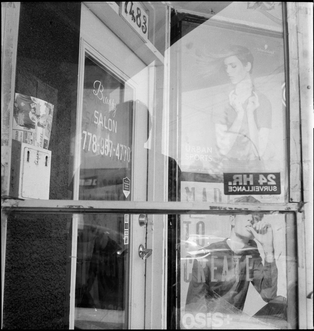 A black and white medium format film photograph of the exterior of a beauty salon at number 1483. The door and window glass reflect the street behind the camera, layering over posters for Schwarzkopf hair products and a 24-hour surveillance sticker. The salon name and phone number are painted on the glass.