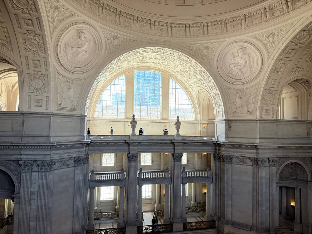 Looking across the upper rotunda of San Francisco City Hall toward a large arched window, with a small wedding party visible on the gallery level being photographed against the elaborate stone and plasterwork