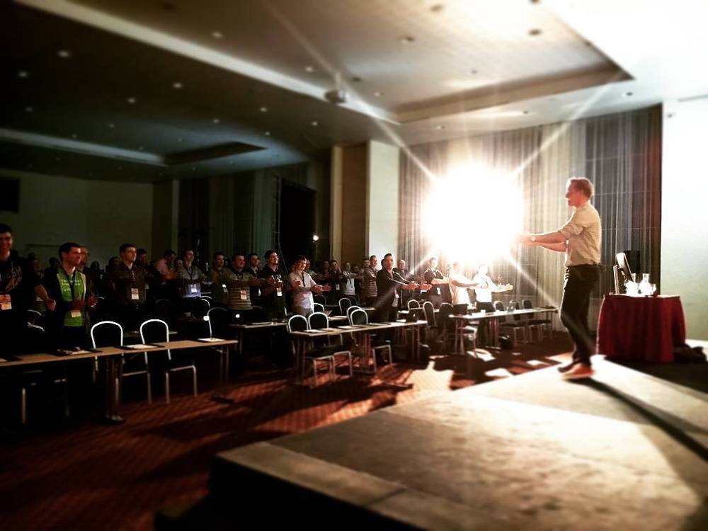 A speaker gesturing on stage in front of a seated conference audience, backlit by bright light.