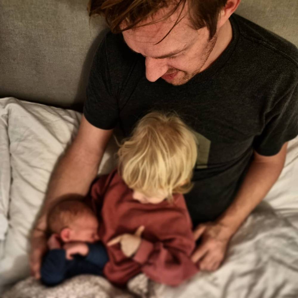 A man sitting on a bed with a toddler beside him and a newborn resting nearby.