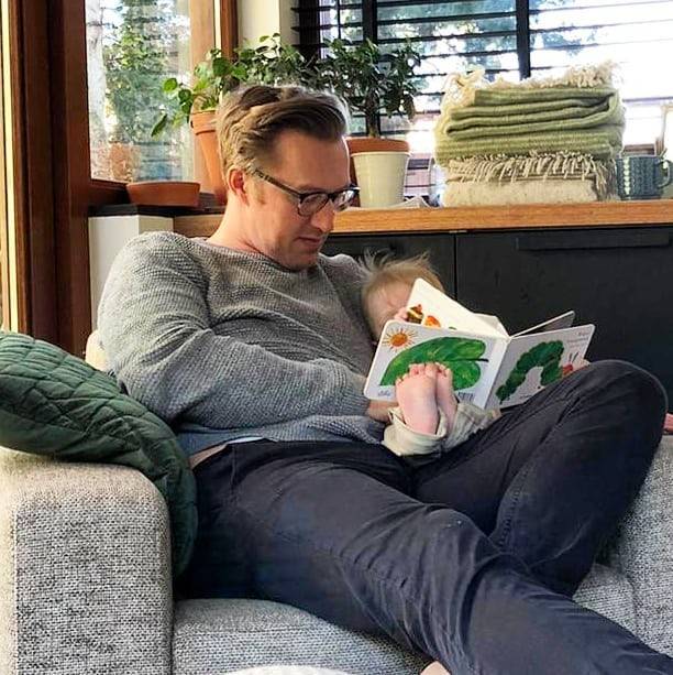 A man sits on a couch reading a picture book to a toddler on his lap.