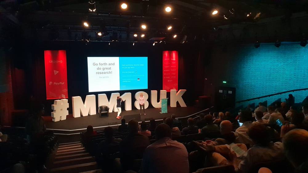 A presenter on a lit stage stands beside a giant MM18UK hashtag sign in front of a seated crowd.