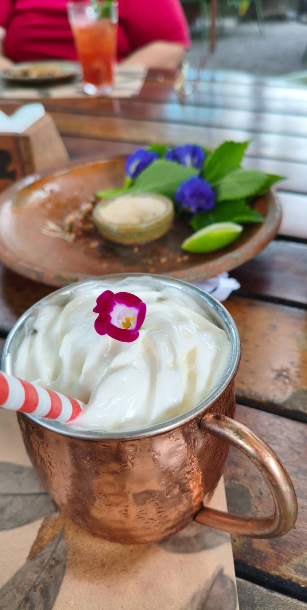 Um drink numa caneca de cobre com bastante creme por cima, decorado com uma flor rosa e um canudo de papel listrado vermelho e branco