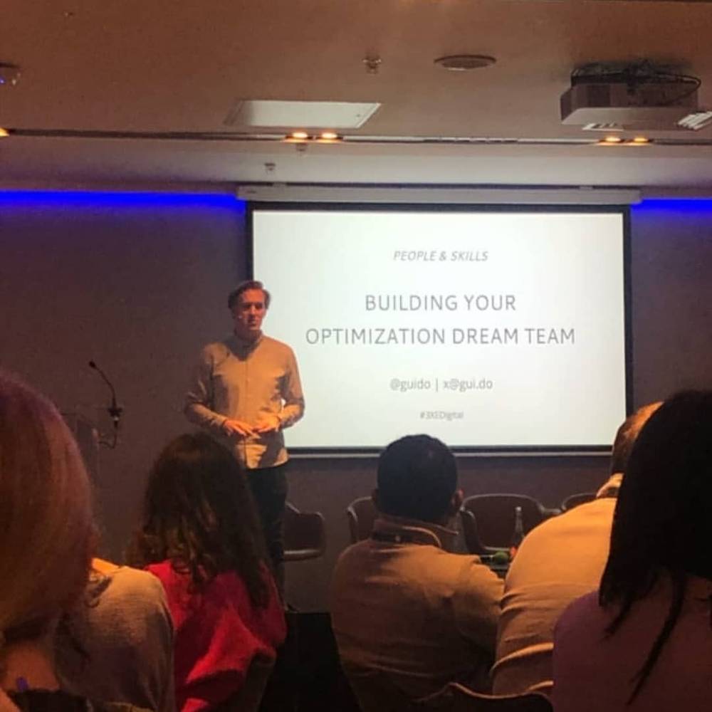A man presents on stage in front of a screen reading "Building Your Optimization Dream Team".